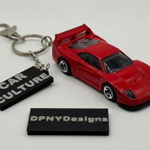 Peut inclure: Une voiture jouet rouge avec des roues noires et un aileron arrière. La voiture fait partie d'un porte-clés avec un fermoir argenté et une étiquette rectangulaire noire qui dit "CAR CULTURE". Une deuxième étiquette noire dit "DPNYDesigns".