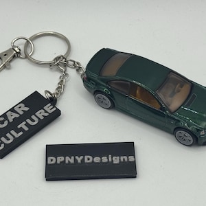May include: A dark green die-cast model car keychain with a silver keyring and clasp. The keychain includes a black rectangular tag with the text "CAR CULTURE" in silver lettering. A second black tag reads "DPNYDesigns".