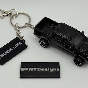 Pode incluir: Chaveiro de caminhonete de brinquedo preta com mosquetão e argola prateados. O chaveiro inclui uma etiqueta preta com o texto "TRUCK LIFE" e outra com "DPNYDesigns".