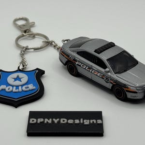 以下が含まれることがあります： 黒いアクセントと側面に「POLICE」の文字がプリントされたシルバーのパトカーのおもちゃ。青と黒の警察バッジのキーホルダーが付いています。「DPNYDesigns」と書かれた黒い長方形のサイン。