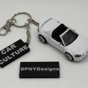 May include: A white toy car with black wheels and a black interior is next to a black rectangular keychain with the text "CAR CULTURE". A second black rectangle with the text "DPNYDesigns" is also present. The keychain has a silver clasp and chain.