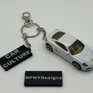 Könnte beinhalten: Ein weißes Spielzeugauto mit goldfarbenen Rädern und einem Schlüsselanhänger mit einem schwarzen Anhänger mit der Aufschrift "CAR CULTURE". Ein zweiter schwarzer Anhänger trägt die Aufschrift "DPNYDesigns".