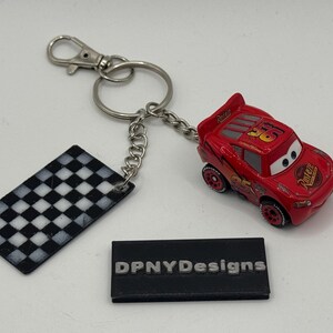 May include: A red Lightning McQueen toy car keychain with a silver chain and clasp. The keychain includes a black and white checkered flag tag. The car has the number 95 on it.