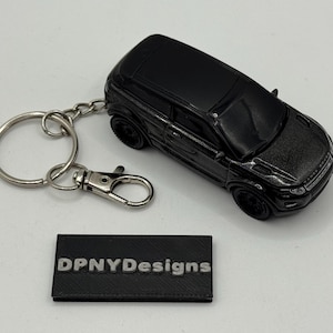 May include: Black die-cast model car keychain. The keychain features a miniature SUV with a silver key ring and clasp. A black rectangular tag with the text "DPNYDesigns" is also included.