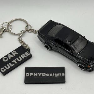 May include: A black die-cast model car keychain with a silver keyring and clasp. The keychain includes a black rectangular tag with the words "CAR CULTURE" in white. A second black tag with "DPNYDesigns" is also present.
