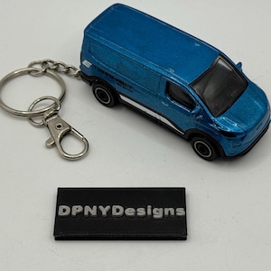 May include: A blue die-cast van keychain with a silver keyring and clasp. The van has black wheels and a white stripe along the side. A black rectangular base with the text "DPNYDesigns" is also present.