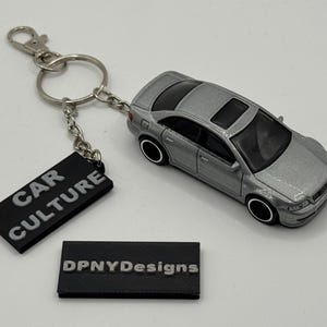 May include: Silver car keyring with a black rectangular tag that reads "CAR CULTURE". The keyring also includes a small black rectangle with the text "DPNYDesigns". The car has black wheels and a sunroof.