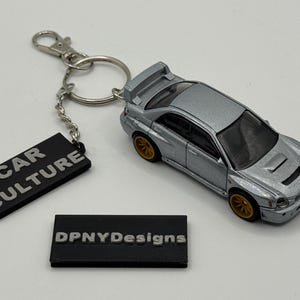 Puede incluir: Un llavero de coche modelo plateado con ruedas doradas y un alerón trasero. El llavero incluye una etiqueta negra con el texto "CAR CULTURE" y otra con "DPNYDesigns".