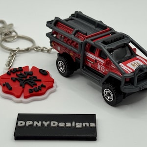 May include: A red and black toy fire truck with a black roll cage, next to a keychain with a red and white emblem. The keychain has the text "Fire Dept" and a silver keyring. A black rectangular block with the text "DPNYDesigns" is also present.