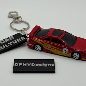 Puede incluir: Un coche de juguete rojo con detalles amarillos y el número 12, sujeto a un llavero con una etiqueta negra que dice "CAR CULTURE". Una segunda etiqueta negra dice "DPNYDesigns".