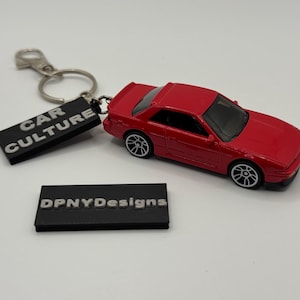 Pode incluir: Um carro de brinquedo vermelho com rodas pretas e um chaveiro com uma etiqueta preta que diz "CAR CULTURE". Uma segunda etiqueta preta diz "DPNYDesigns".