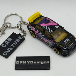 May include: A black Subaru rally car toy keychain with a yellow rear spoiler, pink and purple geometric graphics, and the number 016. Attached is a black rectangular tag with the white text CAR CULTURE. A separate black tag reads DPNYDesigns.
