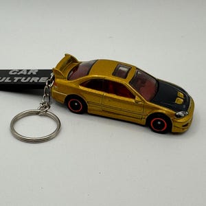 May include: A gold-coloured toy car keyring with a black bonnet and red detailing. The keyring has a silver ring and a black rectangular tag that reads "CAR CULTURE". The car features a spoiler and a sunroof.
