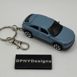 May include: A blue die-cast car keyring with a silver ring and clasp. The car has black wheels and trim. A black rectangular tag with the text "DPNYDesigns" is also present.