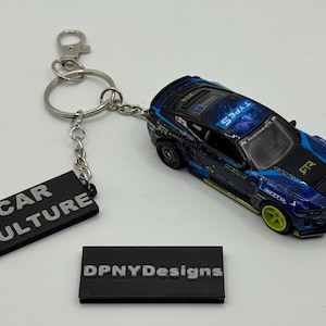 Op de afbeelding: Een blauwe en zwarte speelgoedautosleutelhanger met een zilveren sleutelring en een zwart rechthoekig label met de tekst "CAR CULTURE". De speelgoedauto heeft gele wielen en "TYPES" staat op het dak. Een tweede zwart label leest "DPNYDesigns".