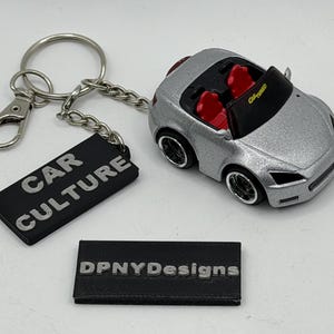 May include: A silver convertible car keychain with red seats and black interior. The windshield features the text Car Tuned. Attached is a black rectangular tag with the embossed text Car Culture. A separate black rectangular tag reads DPNYDesigns.