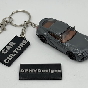 Pode incluir: Um porta-chaves de carro desportivo cinzento com um anel de metal e uma etiqueta retangular preta com o texto branco CAR CULTURE. O modelo de carro tem rodas pretas e um interior castanho. Uma placa retangular preta com DPNYDesigns.