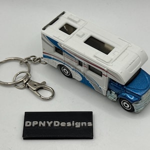 May include: A white and blue toy RV keychain with a silver key ring and clasp. The RV has blue and silver wave-like graphics on the side and a blue front. A black rectangular tag with the text "DPNYDesigns" is below the keychain.