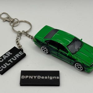 May include: Green toy car keyring with a silver chain and clasp. The keyring includes a black rectangular tag with the words "CAR CULTURE" in white. A second black tag displays "DPNYDesigns".