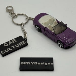 May include: A purple convertible car keyring with a silver chain and a black tag that reads "CAR CULTURE". The keyring also includes a black tag with the text "DPNYDesigns". The car has white interior.
