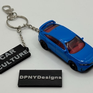 May include: A blue toy car keychain with a silver chain and clasp. The car has a red interior. A black rectangular tag with the words "CAR CULTURE" is attached. Another tag reads "DPNYDesigns".