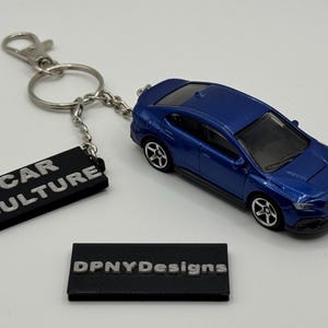 May include: A blue die-cast model car keychain with a silver keyring and a black tag that reads "CAR CULTURE". A second black tag reads "DPNYDesigns".