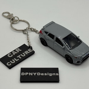 Könnte beinhalten: Ein grauer Druckguss-Autoschlüsselanhänger mit einem schwarzen rechteckigen Anhänger mit der weißen Aufschrift "CAR CULTURE". Der Schlüsselanhänger enthält auch ein kleines schwarzes Etikett mit "DPNYDesigns" in Weiß. Das Auto ist ein Miniaturmodell.