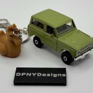 Peut inclure: Un SUV jouet vert olive avec des roues noires est à côté d'un porte-clés cheval marron. Le porte-clés est attaché à un anneau argenté. Un panneau rectangulaire noir avec le texte "DPNYDesigns" est en bas.
