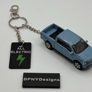 May include: A light blue toy pickup truck with black wheels and a black bed. A black keychain tag with the words "ALL ELECTRIC" and a green lightning bolt is attached. The keychain is connected to a silver keyring.