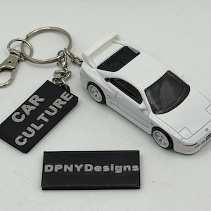 Puede incluir: Un llavero de coche de juguete Toyota MR2 blanco con una anilla de metal y una etiqueta rectangular negra con el texto en relieve blanco CAR CULTURE. Una etiqueta rectangular negra separada con el texto en relieve blanco DPNYDesigns se sitúa debajo.