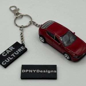 Puede incluir: Un llavero de coche de juguete rojo con una cadena plateada y una etiqueta negra que dice "CAR CULTURE". Una segunda etiqueta negra dice "DPNYDesigns". El coche es un modelo pequeño y detallado.