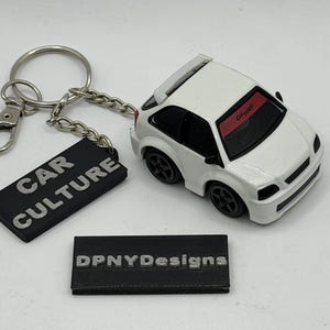 May include: A white toy car keychain with a rear spoiler and black wheels. A metal ring and chain attach to a black rectangular tag with the white text CAR CULTURE. A separate black rectangular tag features the white text DPNYDesigns.