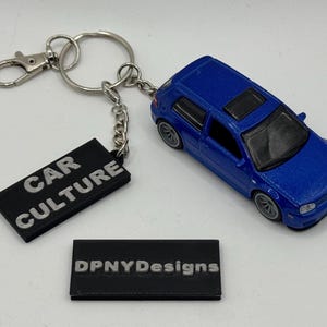 Könnte beinhalten: Ein blauer Druckguss-Auto-Schlüsselanhänger mit einem silbernen Schlüsselring und Verschluss. Der Schlüsselanhänger enthält ein schwarzes rechteckiges Etikett mit dem Text "CAR CULTURE". Ein zweites schwarzes Etikett mit dem Text "DPNYDesigns" ist ebenfalls vorhanden.