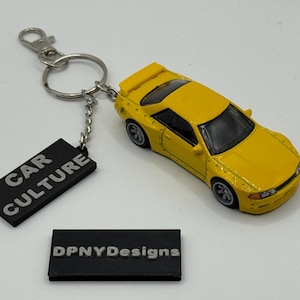May include: Yellow toy car keychain with a black tag that reads "CAR CULTURE". The keychain also includes a small black rectangle with the text "DPNYDesigns".