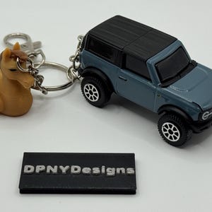 Puede incluir: Un SUV de juguete azul grisáceo con ruedas negras y techo negro está unido a un llavero con un dije en forma de caballo. El llavero también incluye un pequeño bloque rectangular negro con el texto "DPNYDesigns".