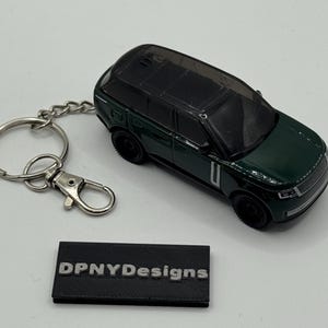 Puede incluir: Un llavero de SUV modelo fundido a presión de color verde oscuro con ruedas negras y un llavero plateado. El llavero incluye un pequeño letrero rectangular negro con el texto "DPNYDesigns" en plata.
