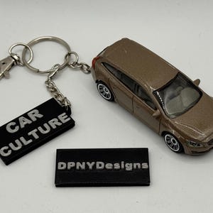 Op de afbeelding: Een bruine modelauto naast een sleutelhanger met een zwart label met de tekst "CAR CULTURE". Een tweede zwart label leest "DPNYDesigns". De auto heeft zwarte wielen en een zilveren voorruit.