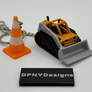 May include: A miniature construction-themed keyring. It features a bright orange and white traffic cone, a yellow and grey mini dozer, and a black rectangular base with the text "DPNYDesigns".