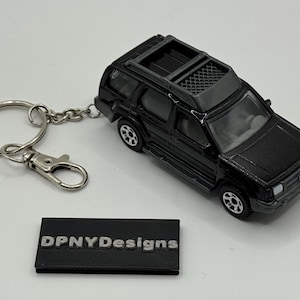Puede incluir: Llavero de SUV negro con un llavero y cierre plateados. El llavero incluye una pequeña etiqueta rectangular negra con el texto "DPNYDesigns" en blanco. El SUV tiene ruedas negras y baca.