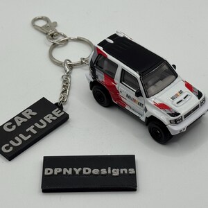 May include: A white and black toy SUV with red detailing, a keychain, and a black tag that reads "CAR CULTURE". The keychain is attached to a silver ring. A black tag with "DPNYDesigns" is also present.