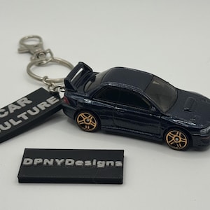 May include: A navy blue die-cast model car keychain with gold-colored wheels and a rear spoiler. The keychain includes a black rectangular tag that reads "CAR CULTURE". A second black tag reads "DPNYDesigns".