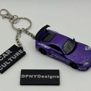 Peut inclure: Une voiture miniature violette avec un aileron noir et des autocollants de course. La voiture est attachée à un porte-clés avec une étiquette noire qui dit "CAR CULTURE". Une deuxième étiquette noire dit "DPNYDesigns".