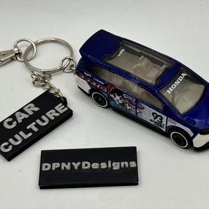 Może przedstawiać: Niebiesko-biały brelok do kluczy z modelem Hondy z czarną etykietą z napisem "CAR CULTURE". Samochód ma przezroczysty dach i grafikę z kreskówek. Druga czarna etykieta głosi "DPNYDesigns".