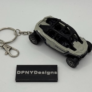 Puede incluir: Un llavero de veh&iacute;culo todoterreno gris y negro en miniatura. El coche de juguete tiene una jaula antivuelco y ruedas detalladas. El llavero incluye una anilla partida y un mosquet&oacute;n. Tambi&eacute;n hay una peque&ntilde;a etiqueta rectangular negra con el texto "DPNYDesigns".
