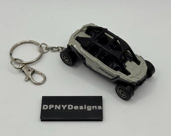 May include: A miniature gray and black off-road vehicle keychain. The toy car has a roll cage and detailed wheels. The keychain includes a split ring and a lobster clasp. A small black rectangular tag with the text "DPNYDesigns" is also present.