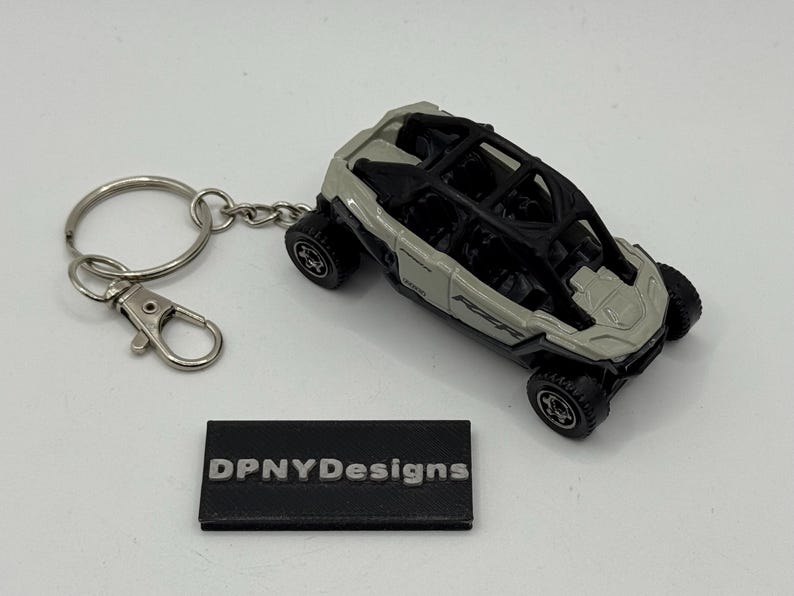 Puede incluir: Un llavero de veh&iacute;culo todoterreno gris y negro en miniatura. El coche de juguete tiene una jaula antivuelco y ruedas detalladas. El llavero incluye una anilla partida y un mosquet&oacute;n. Tambi&eacute;n hay una peque&ntilde;a etiqueta rectangular negra con el texto "DPNYDesigns".