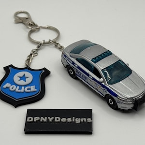 May include: Silver police car toy with blue and white detailing, alongside a blue and black police badge keyring. The keyring has a silver clasp. A black rectangular block with the text "DPNYDesigns" is also present.