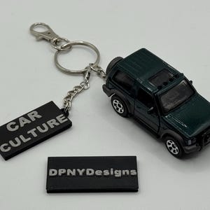 以下が含まれることがあります： シルバーのキーリングとチェーンが付いた、濃い緑色の玩具SUVキーホルダー。「CAR CULTURE」の文字が銀色の黒い長方形のタグと「DPNYDesigns」と書かれた2つ目の黒いタグが付いています。