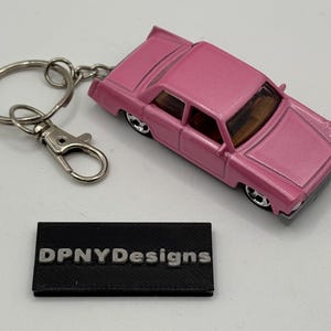 Może przedstawiać: Różowy brelok do kluczy w kształcie zabawkowego samochodu ze srebrnym kółkiem i zapięciem. Samochód ma klasyczny design sedana. Obecny jest również mały czarny prostokąt z napisem "DPNYDesigns".