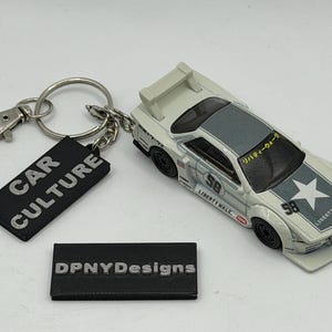 May include: A white toy car keychain featuring a Liberty Walk design with the number 58, a star graphic, and Japanese text on the windshield. Attached is a black rectangular tag with the text CAR CULTURE and a separate black tag labeled DPNYDesigns.